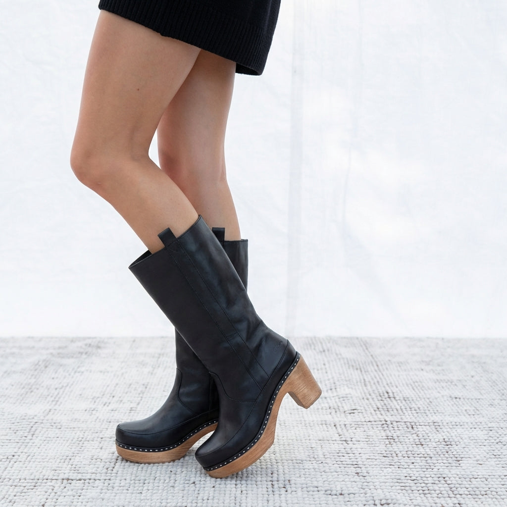 Black leather clog boots with wooden soles worn by a person on a white background
