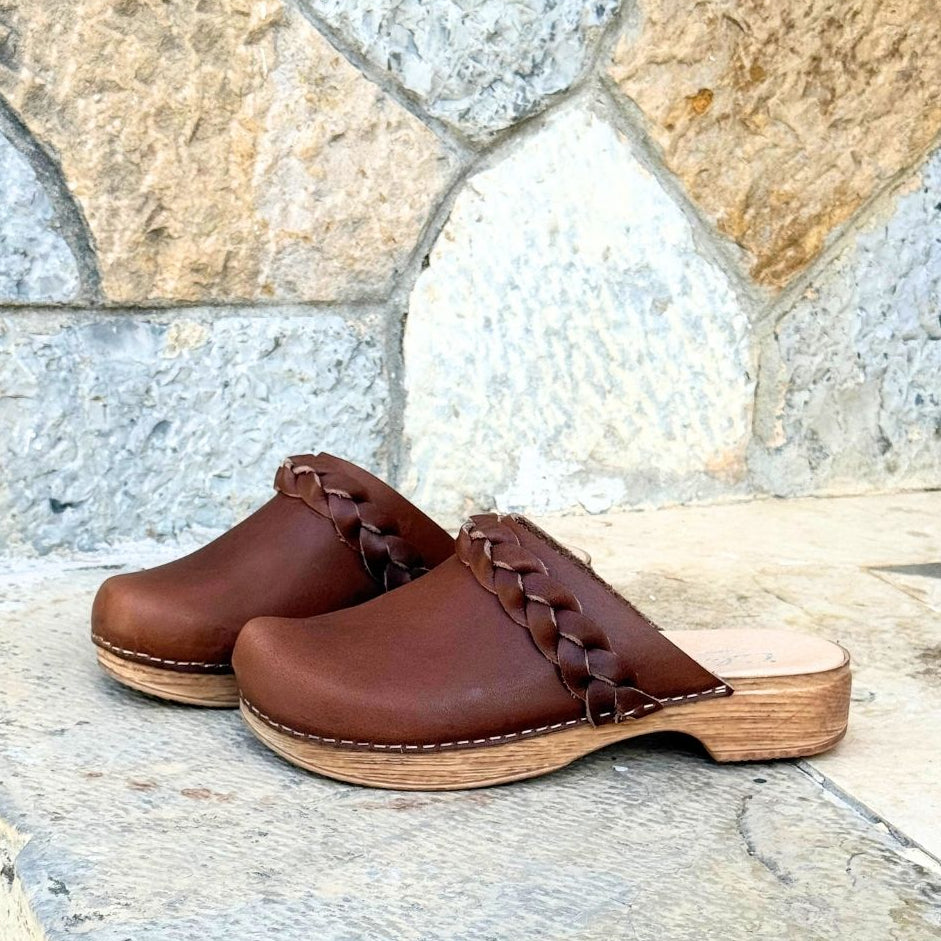 Pair of brown wood-look sole clogs with braided details on a stone surface.