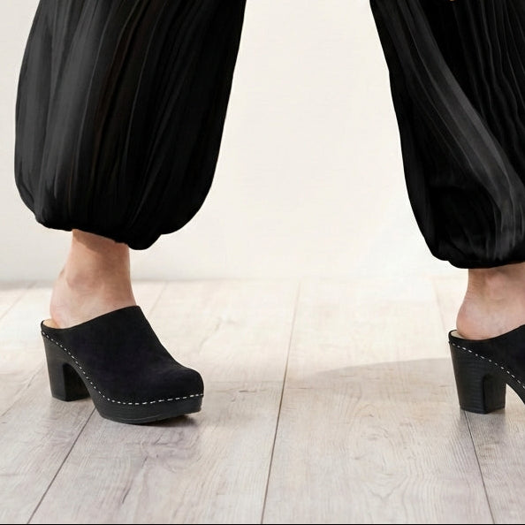Black clogs worn with black pants on a light wooden floor.