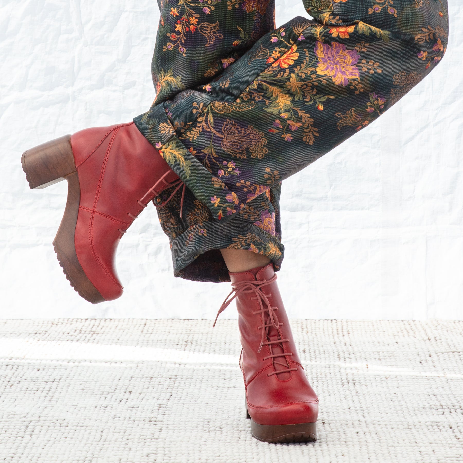Red boots with brown soles worn with  floral patterned pants on a white textured surface.